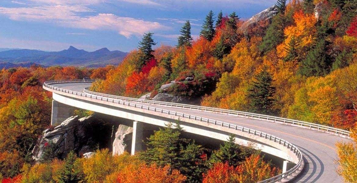 Curved concrete bridge over a rocky gorge, surrounded by vibrant fall foliage and distant mountains.